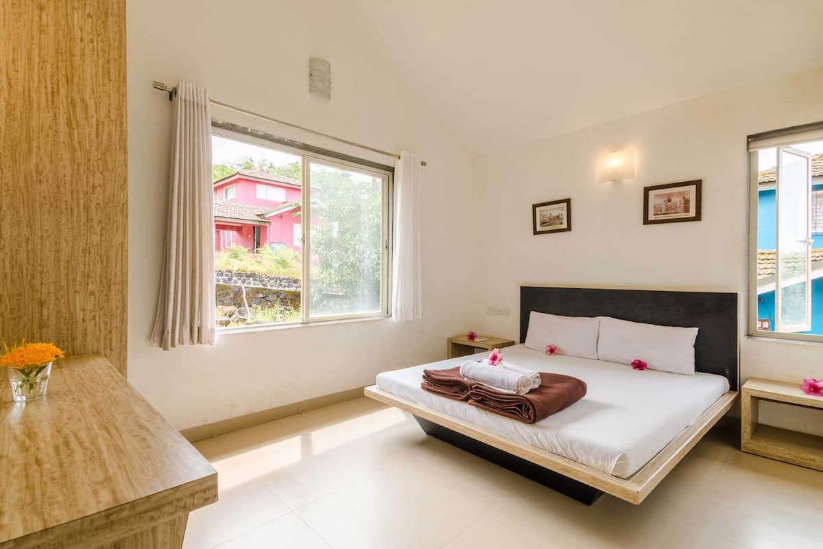 A bedroom with a bed, a nightstand, a window with curtains, a wooden desk, and a vase with flowers.