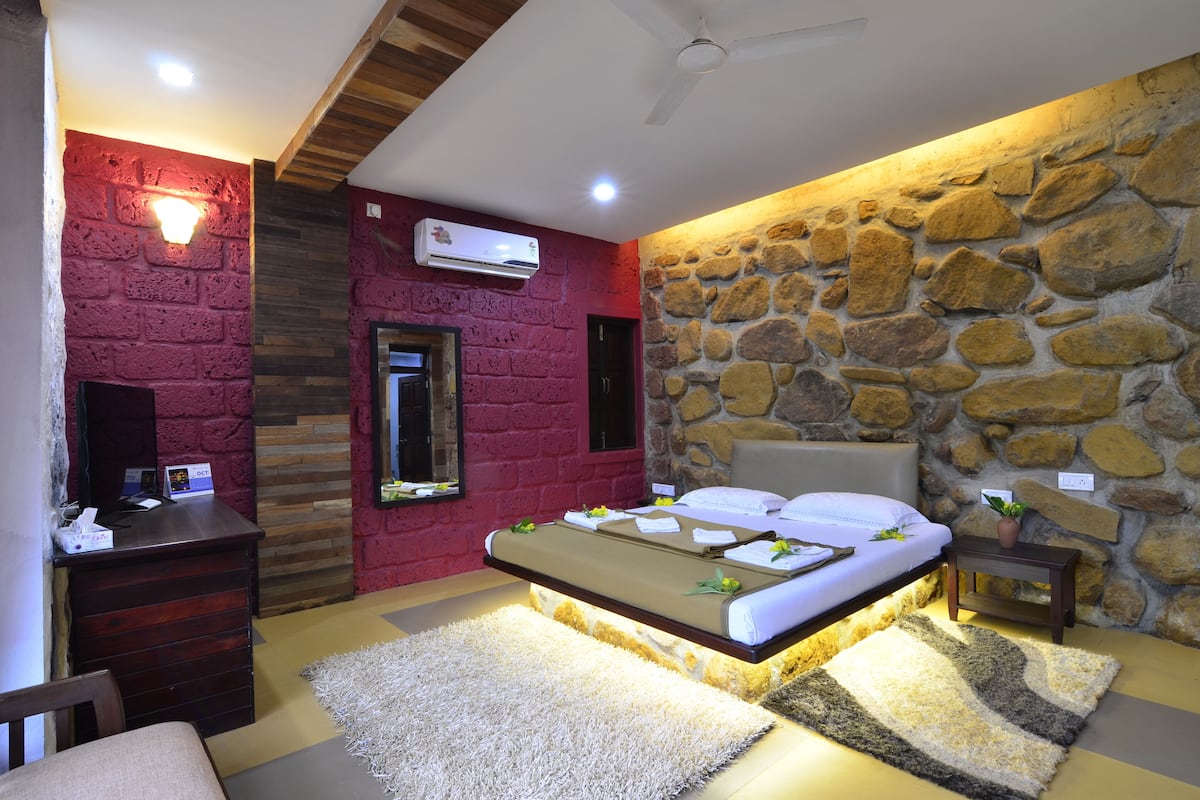 A bedroom with a stone wall, a bed with a headboard, a desk, and an air conditioning unit.