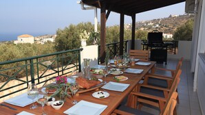 Una mesa de madera dispuesta para una comida, con vista al mar y colinas al fondo.