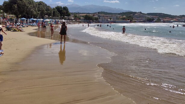 Una playa con gente caminando y nadando, sombrillas y montañas al fondo.