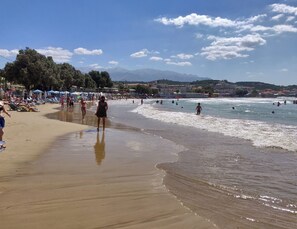 Ein Strand mit Menschen, die spazieren gehen und schwimmen, Sonnenschirmen und einer Berglandschaft im Hintergrund.