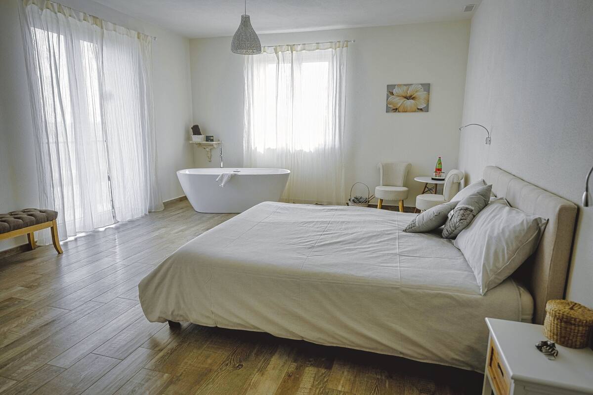 A bedroom with a large bed, a bathtub, a wooden bench, and a small table with a bottle.