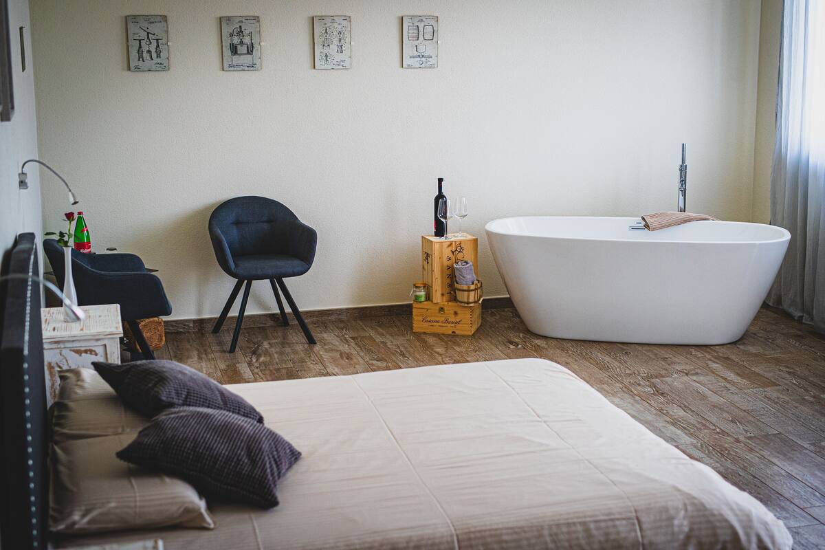 A modern bedroom with a large bed, a wooden stool, a chair, a bathtub, and artwork on the wall.