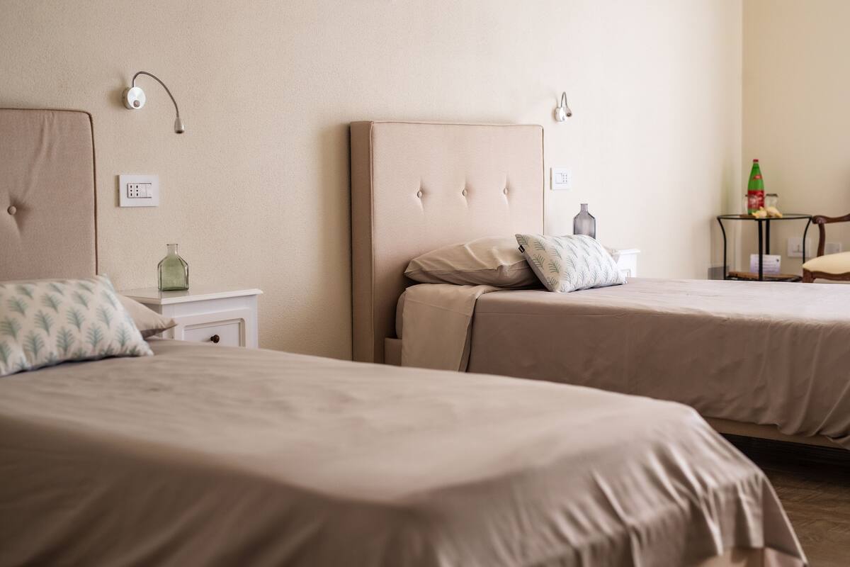 A hotel room with two beds, a nightstand with a green bottle, a vase, and a chair.