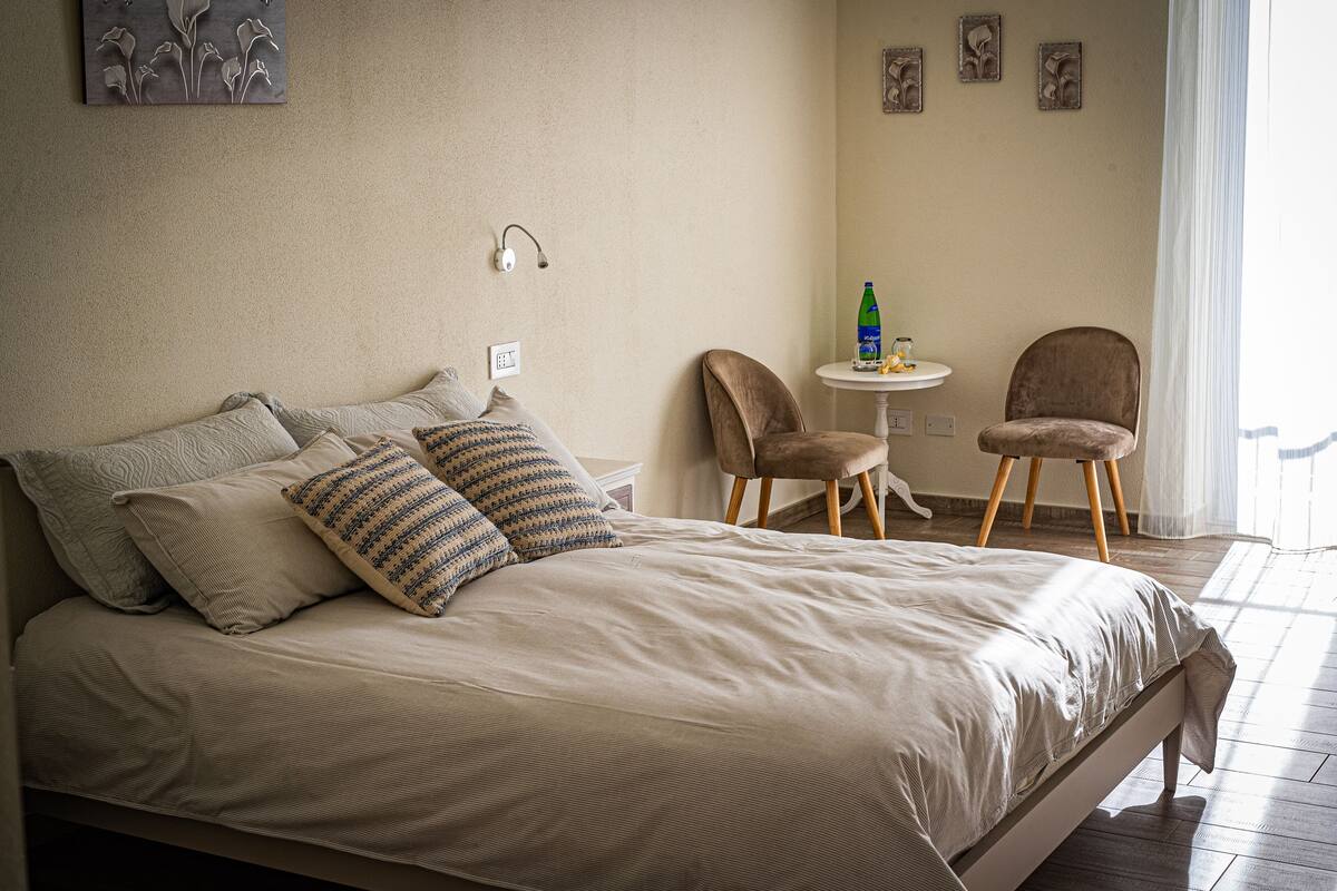 A bedroom with a bed, two chairs, a small table with a bottle and glasses, and wall art.