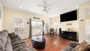 A living room with a sofa, a round coffee table, a TV mounted on the wall, and a dining area with a table and chairs. - 4BR Townhouse with Garage & Pool in Downtown OC- Close to Beach & Boardwalk! (Ocean City)