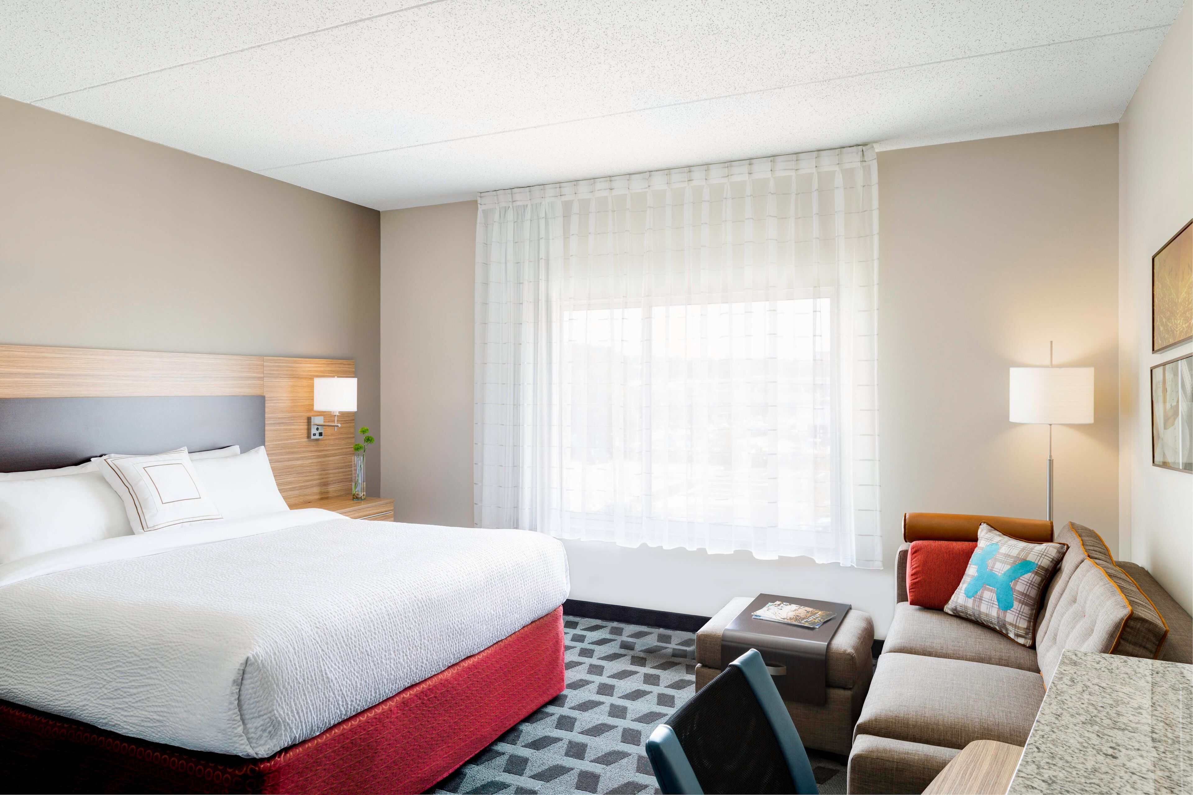 A hotel room with a large bed, a sofa, a small table, and a window with sheer curtains.