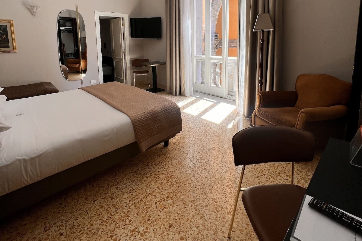 A hotel room with a large bed, a desk, a chair, a TV, and a view of the outside.