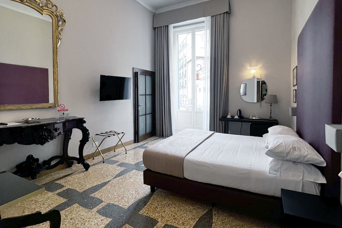 A hotel room with a bed, a desk, a chair, a TV, a mirror, and a window with curtains.