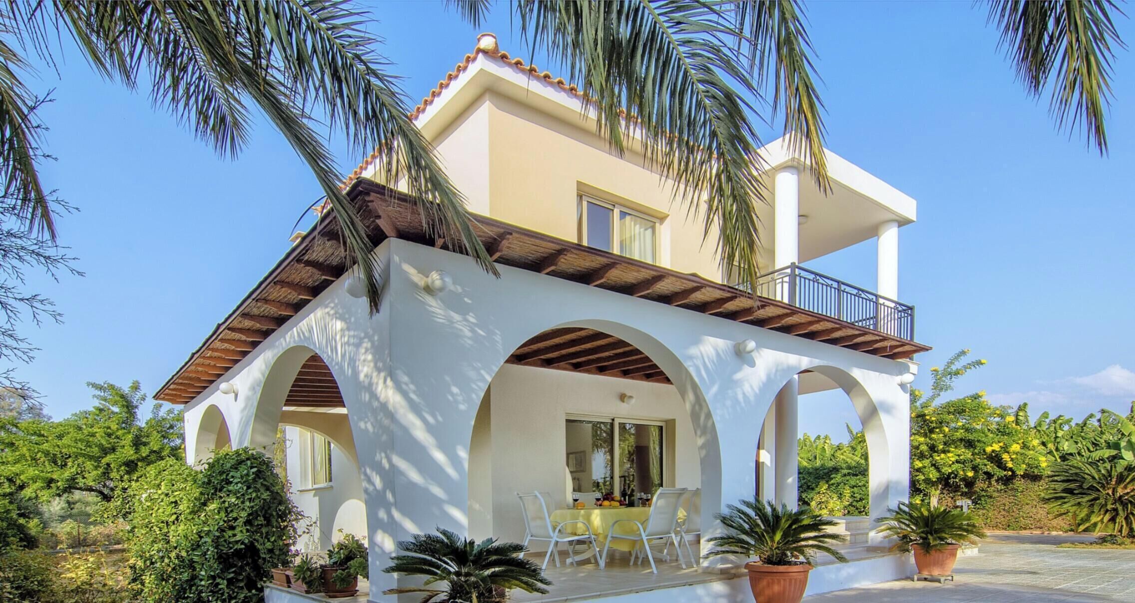 Una villa de dos pisos con un patio cubierto, mobiliario exterior y plantas en macetas.