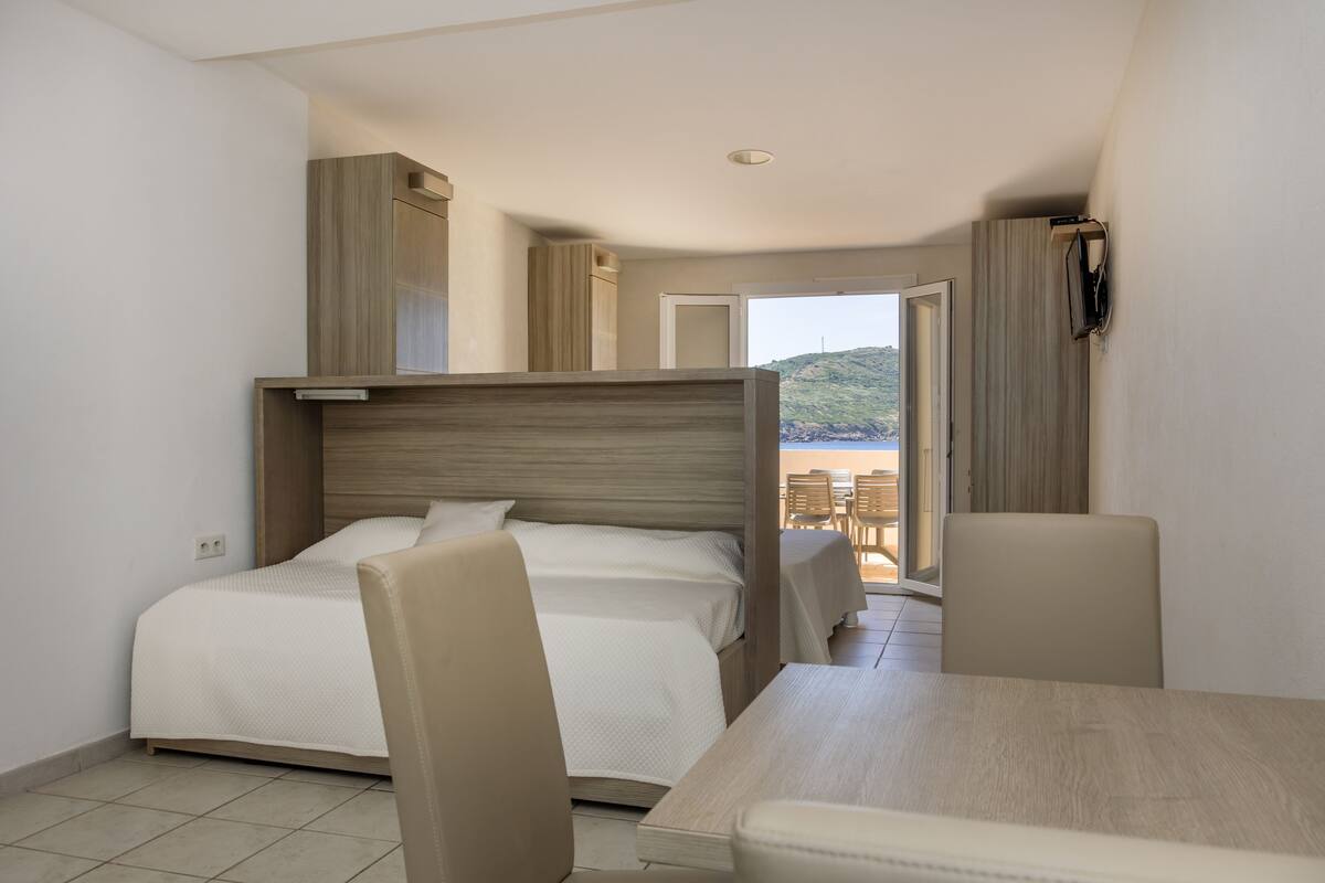 Een hotelkamer met een bed, een tafel, stoelen en uitzicht op het strand door een geopende deur.