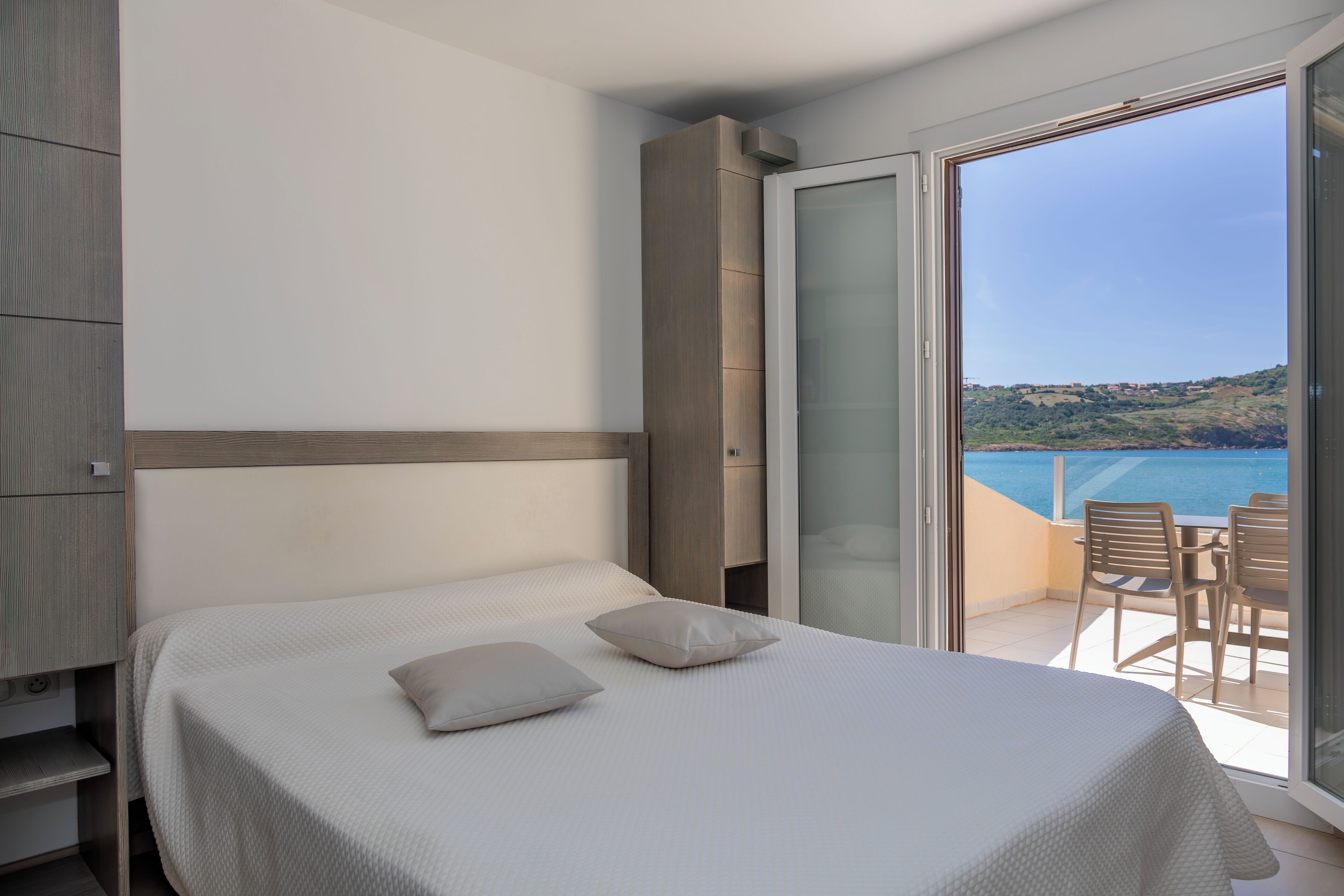 A bedroom with a bed, two pillows, a nightstand, a wardrobe, and a balcony with a table and chairs.