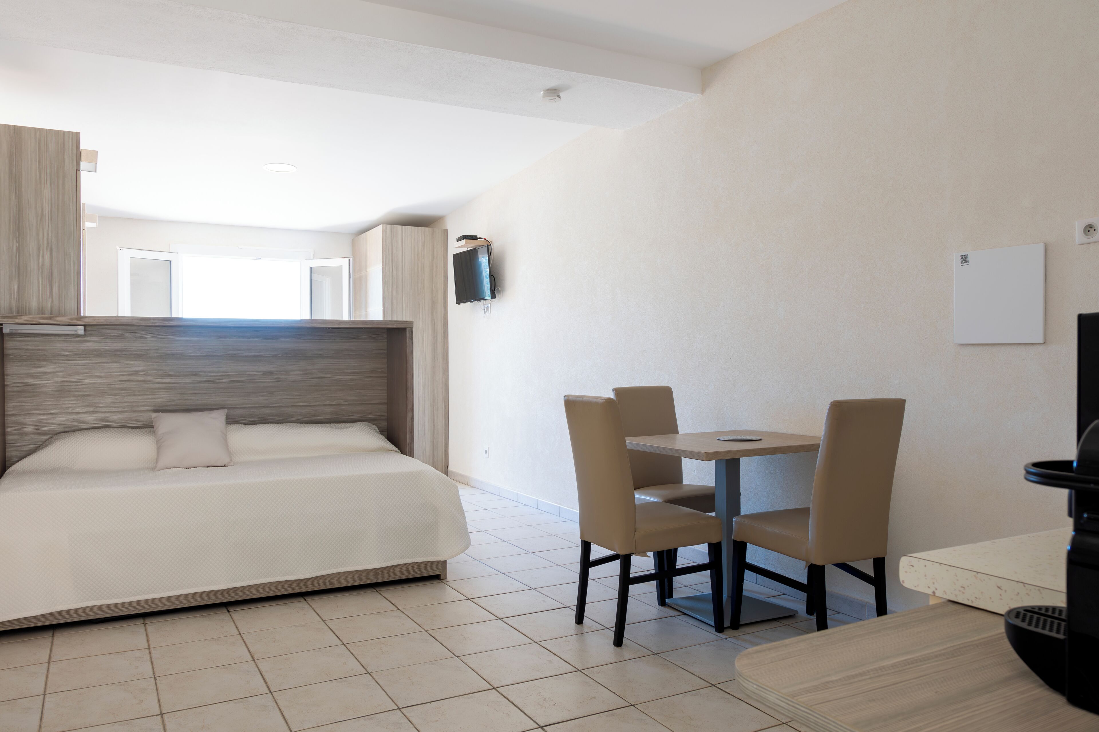 A modern hotel room with a bed, a small dining table, and chairs. There is a TV mounted on the wall and a window providing natural light.
