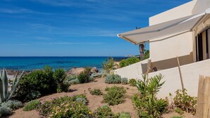 Une propriété située sur la côte, dotée d’une terrasse donnant sur la mer, entourée de diverses plantes et bordée d’un bâtiment blanc.