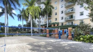Entrada de hotel con suelo de arena, palmeras y un edificio de varios pisos con balcones.