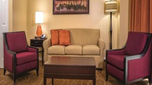Habitación de hotel con un sofá color beige, dos sillones rojos, una mesa de centro de madera, una lámpara y un cuadro en la pared.