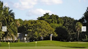 Un campo de golf con una bandera en el green, palmeras y un edificio al fondo.