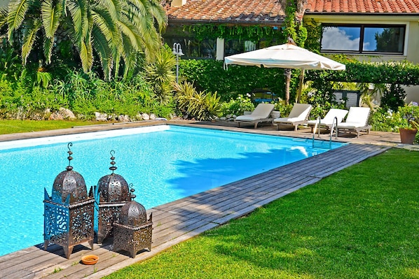 Una piscina con cubierta de madera, rodeada de exuberante vegetación y una casa con tejado de baldosas.