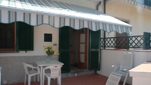 A patio with a table and chairs, a vase with flowers, and a building with green shutters and a striped awning. - Apartment The triangle near the center and the beach (Marina di Campo)