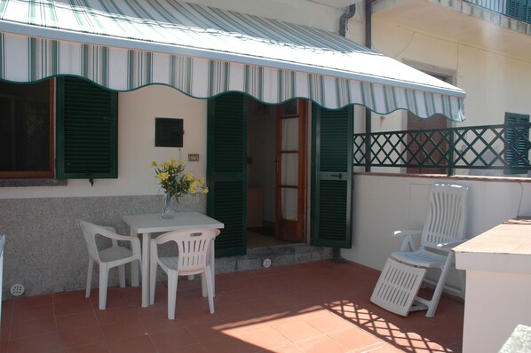 Un patio con una mesa y sillas, un jarrón con flores y un edificio con contraventanas verdes y un toldo a rayas.