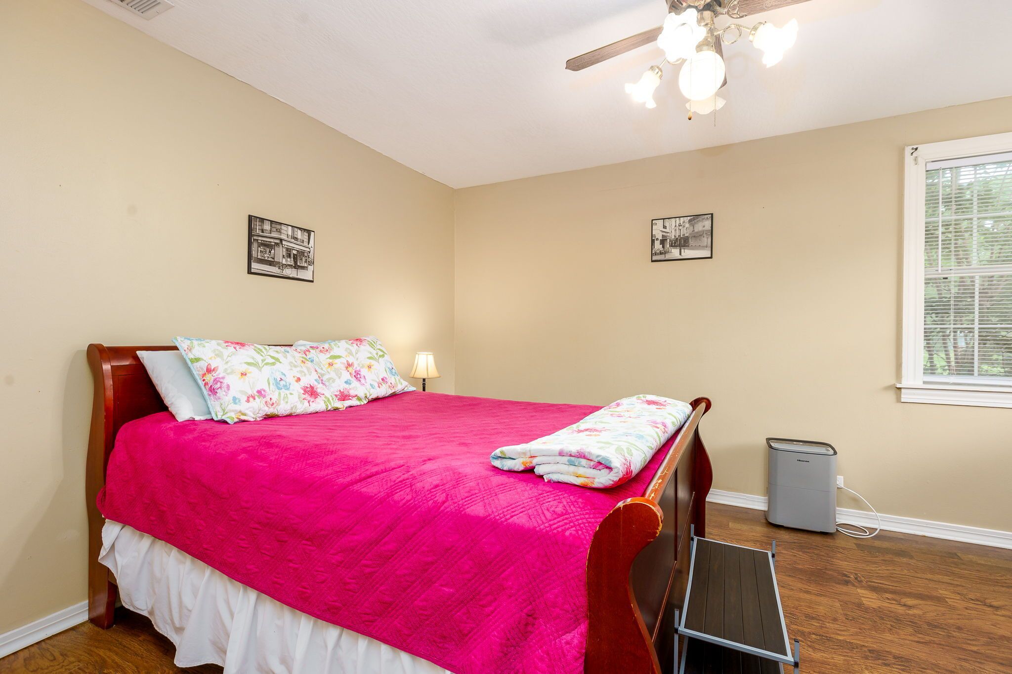 A bedroom with a wooden bed, a pink bedspread, two pillows, a nightstand with a lamp, a ceiling fan, and a window with blinds.