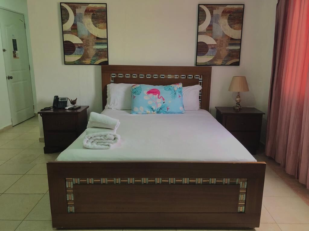 Un lit bien fait, agrémenté d’un coussin à motifs floraux, une table de chevet avec une lampe et des œuvres d’art accrochées aux murs.