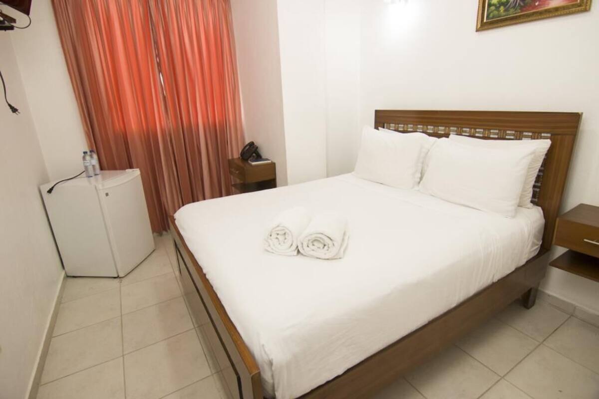 Una habitación de hotel con una cama, una mesita de noche, un armario blanco y una cortina.