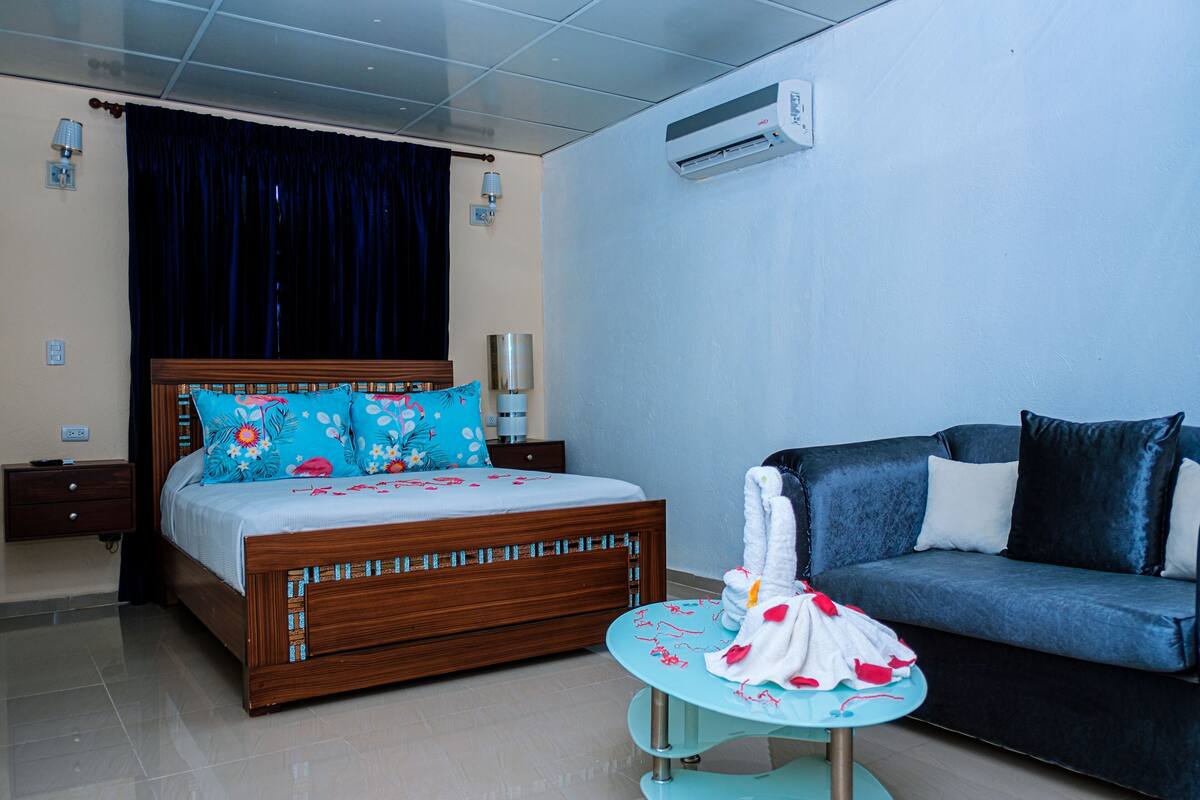 Habitación de hotel con una cama de madera, un sofá azul, una mesa redonda con una toalla y un equipo de aire acondicionado en la pared.
