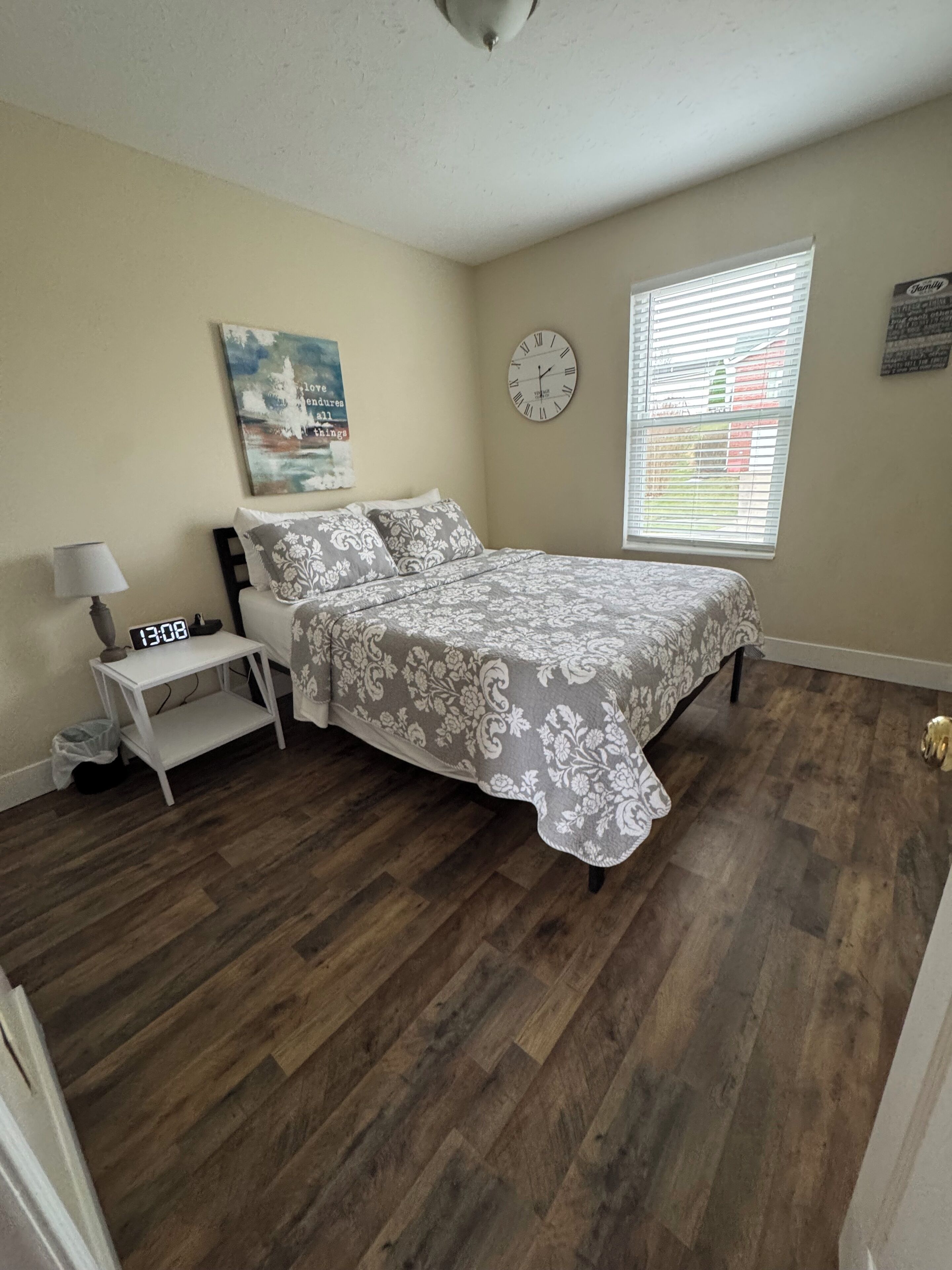 A bedroom with a bed, a bedside table, a wall clock, a window with blinds, and a painting on the wall.
