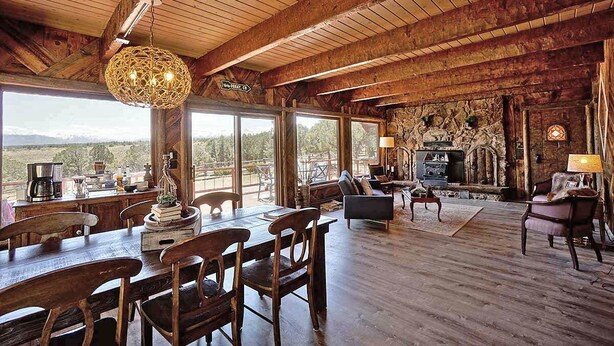 A rustic interior with a wooden dining area, large windows offering a scenic view, and a stone fireplace. - Charming Mountain Chalet with Stunning Views (Montrose)