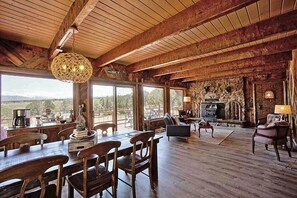 A rustic interior with a wooden dining area, large windows offering a scenic view, and a stone fireplace.