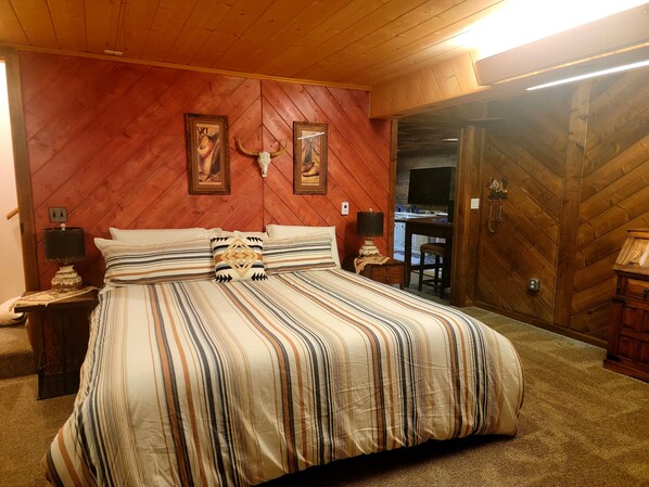 A bedroom with a wooden herringbone ceiling, a bed with striped bedding, two wall-mounted lamps, and a wooden door leading to another room.
