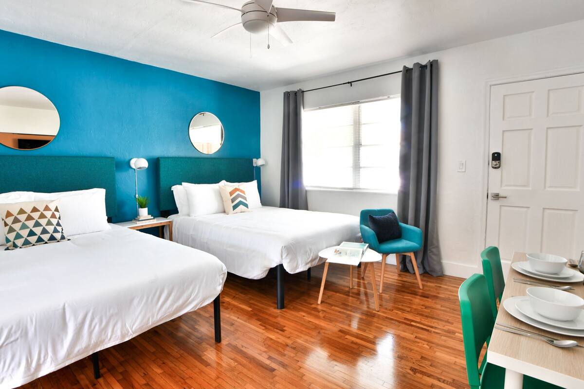 Een hotelkamer met twee bedden, een plafondventilator, een raam met gordijnen, een zithoek met een tafel en stoelen, en een deur.