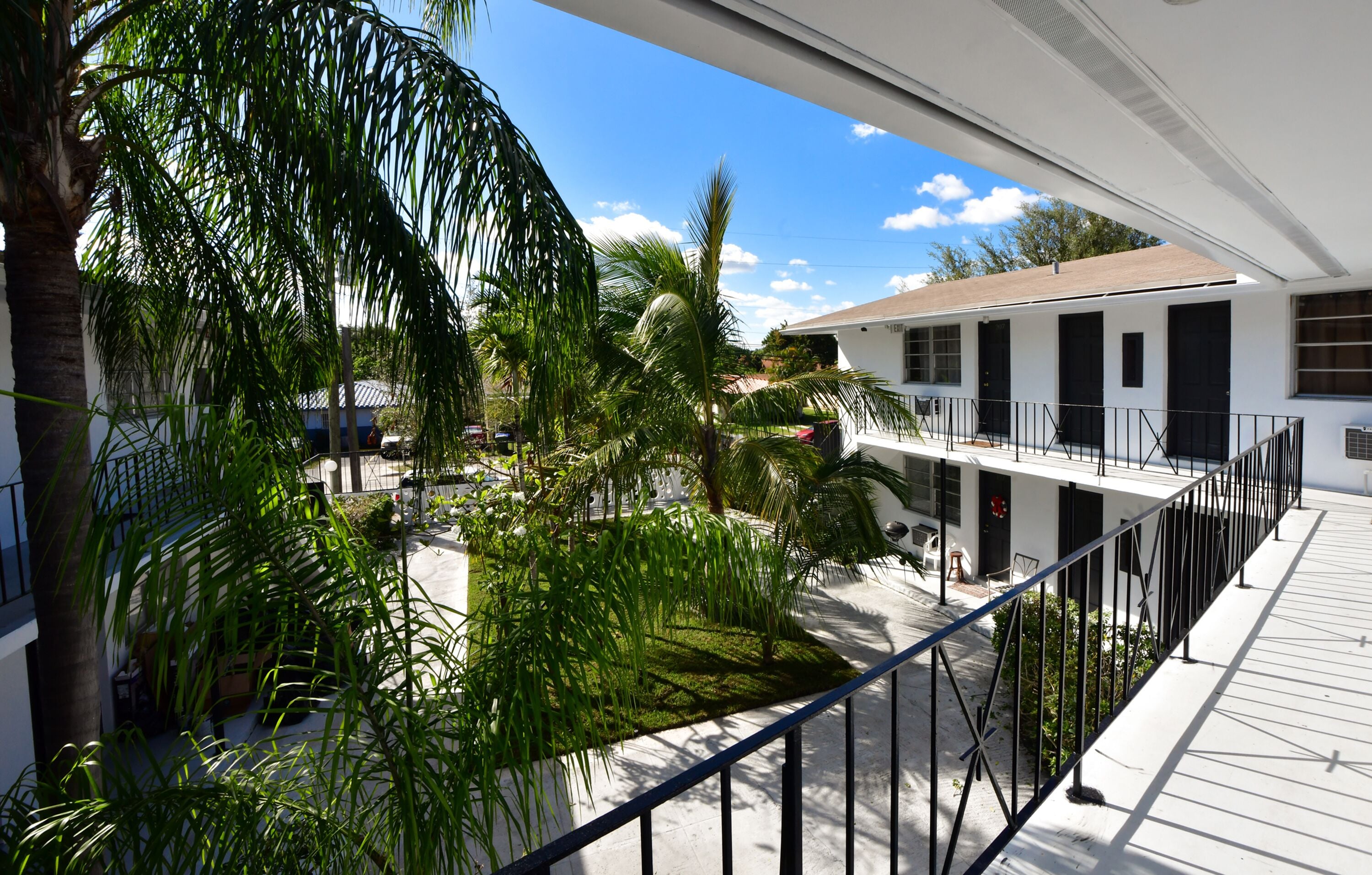 A balcony with a view of a courtyard, palm trees, and a building with multiple windows and doors.