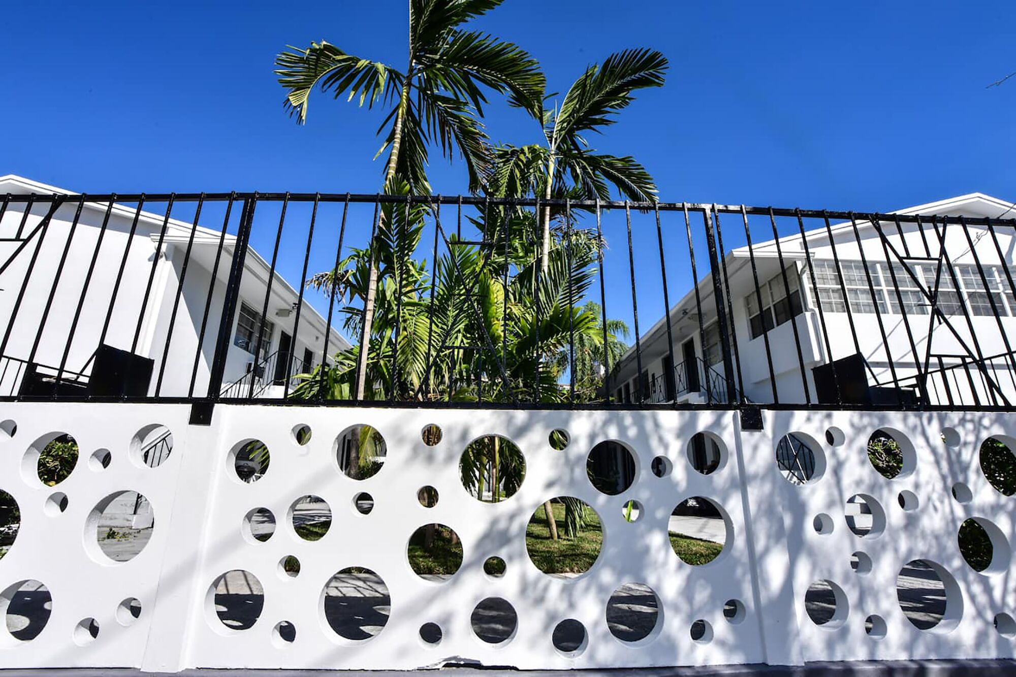 A white fence with circular cutouts reflecting outdoor scenes, a black metal gate, and a clear blue sky.