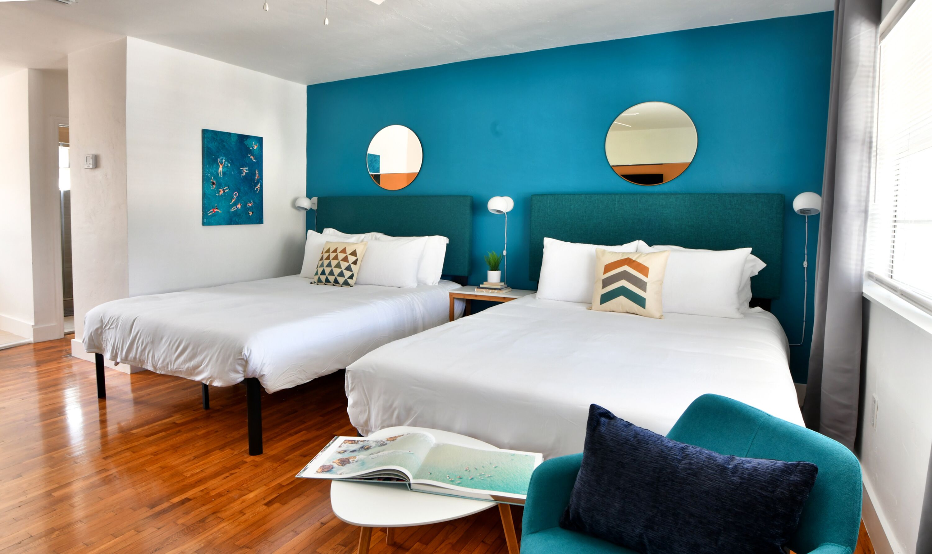 A hotel room with two beds, a teal accent wall, and a small seating area with a magazine.