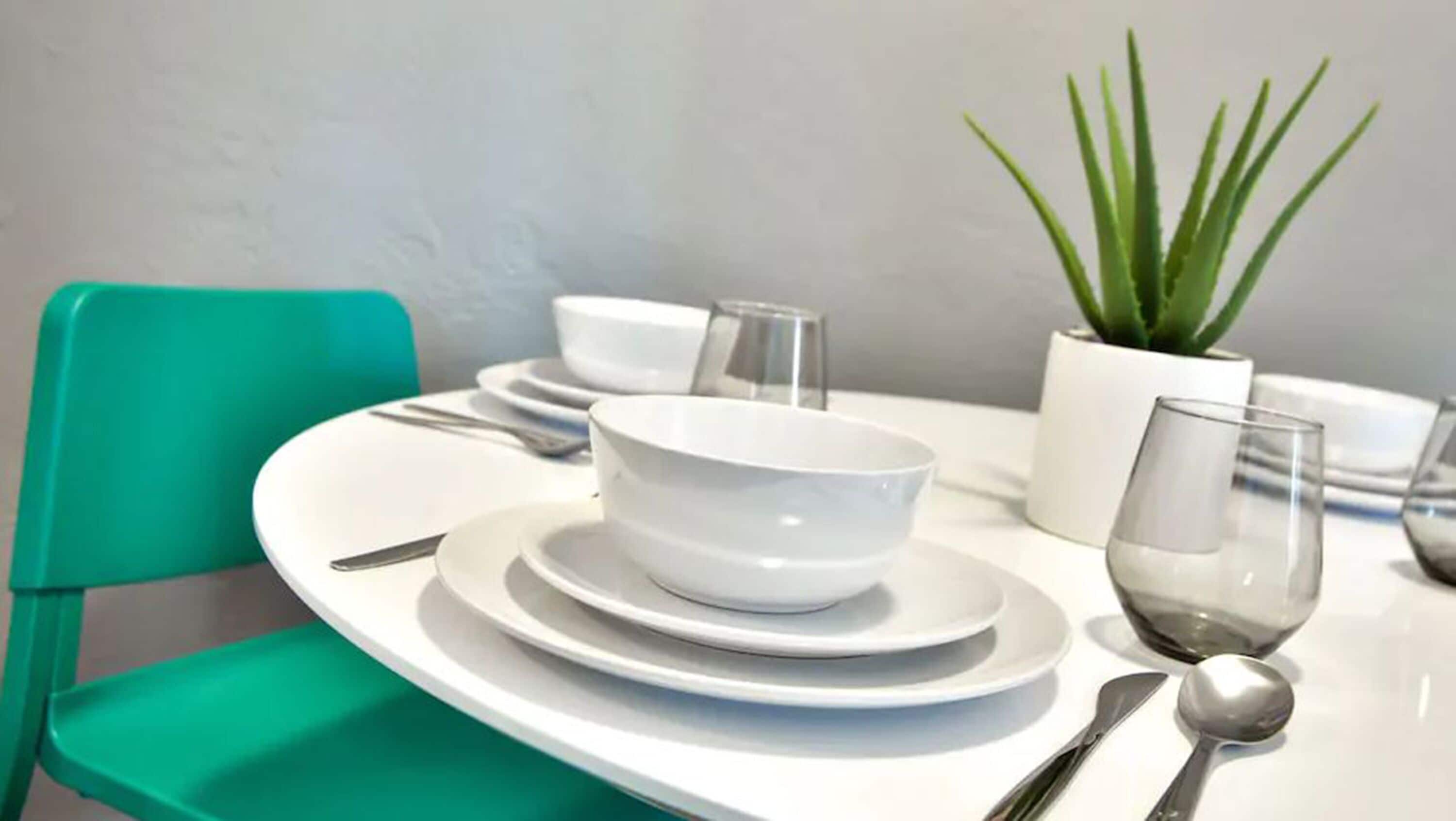 Une table à manger avec de l’assiette, des bols et des couverts blancs, une chaise verte et une plante en pot.