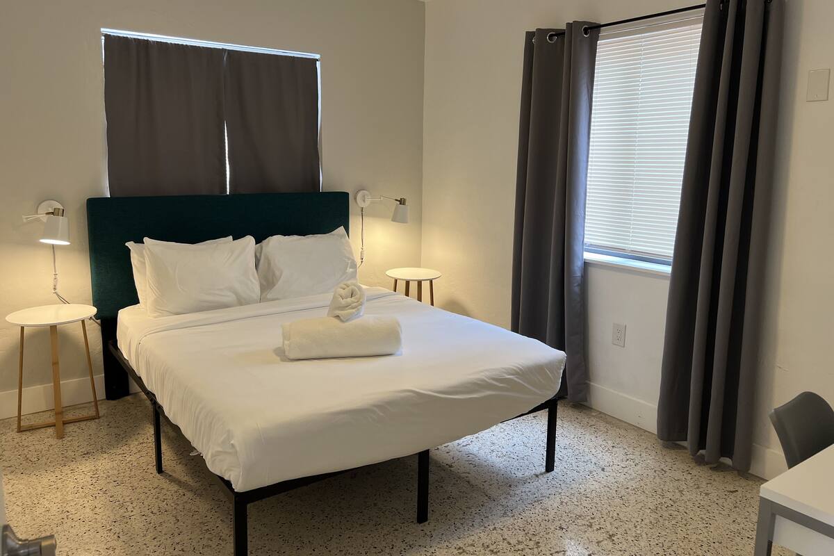 Een netjes opgemaakt bed met witte lakens en een opgevouwen handdoek, een donkergroen hoofdbord, twee bedlampjes, een klein rond tafeltje en een raam met gordijnen.