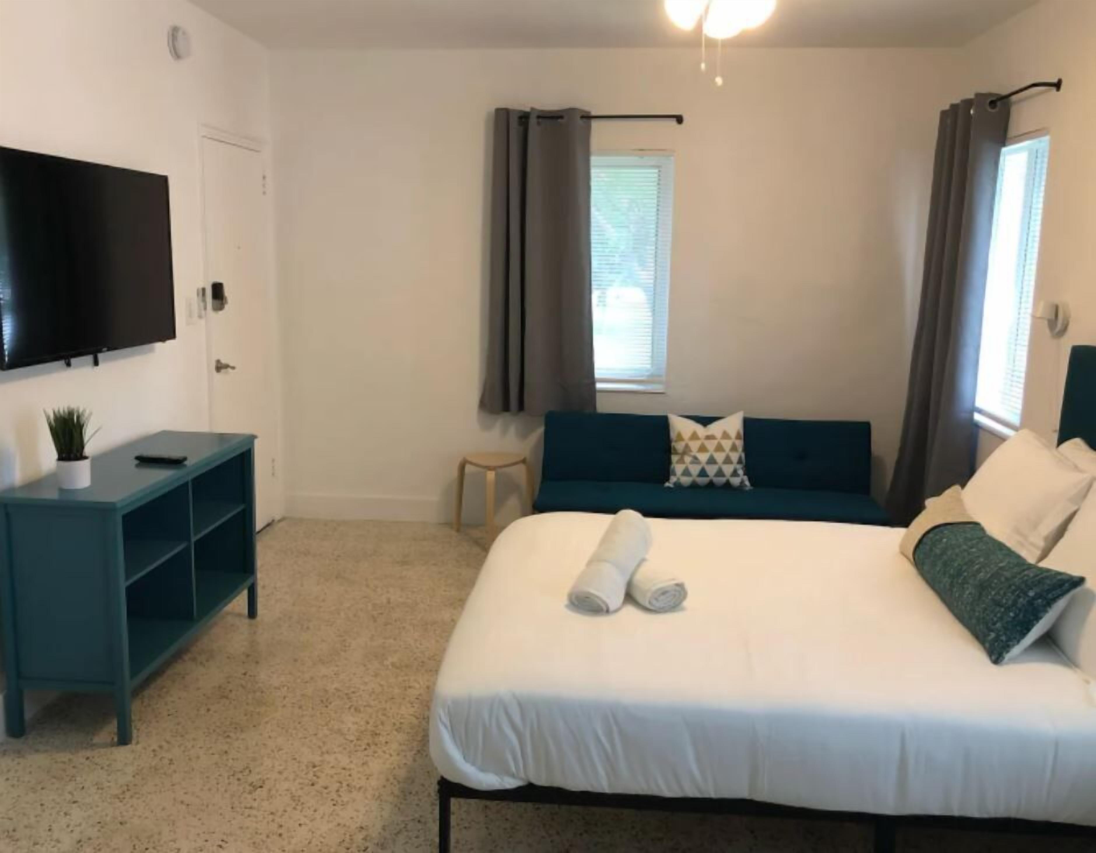 Un dormitorio con cama, televisor, una mesita con planta y ventana con cortinas.