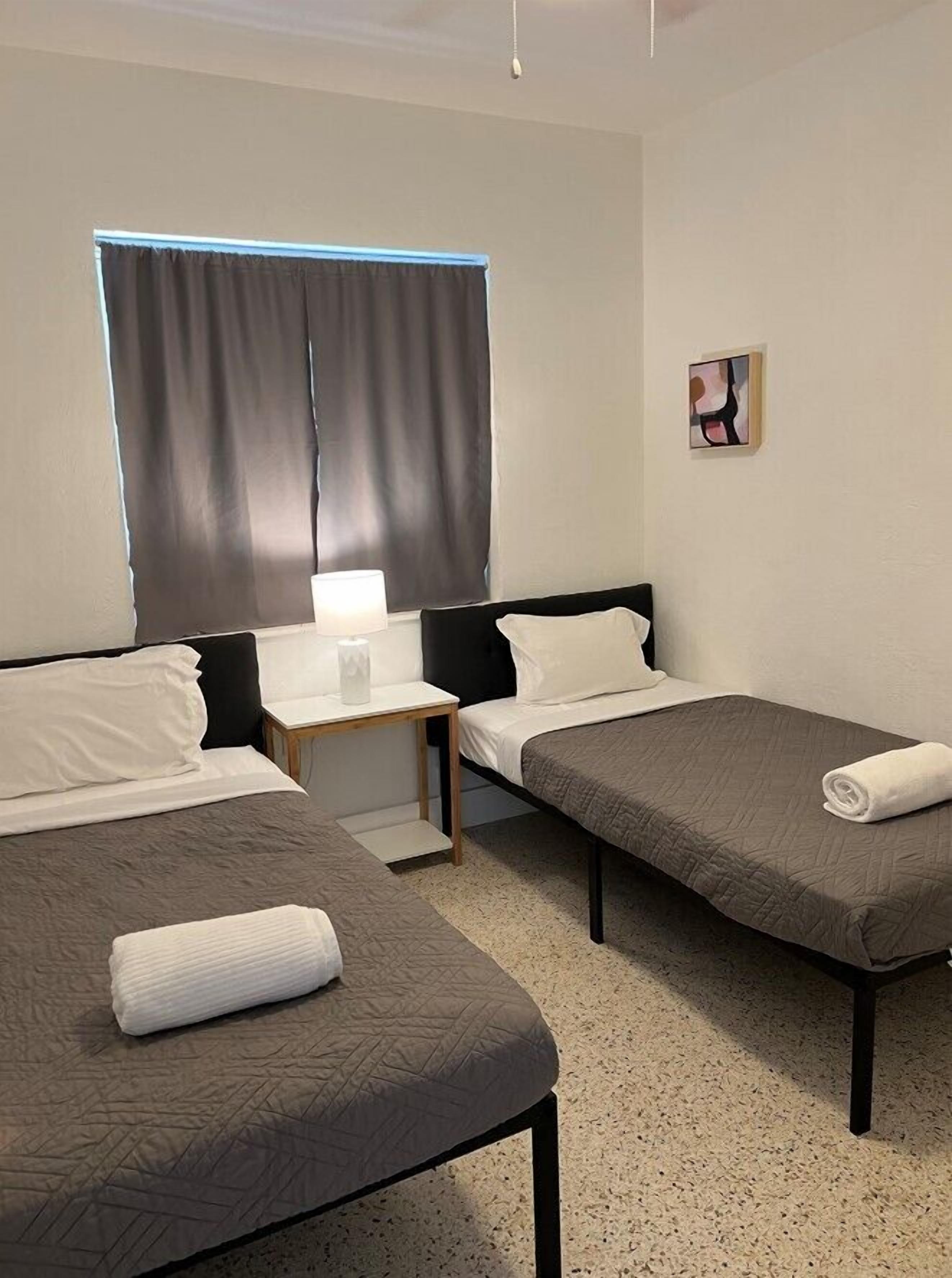 Una habitación de hotel con dos camas individuales, una mesita de noche con lámpara, una ventana con cortinas y una obra de arte colgada en la pared.