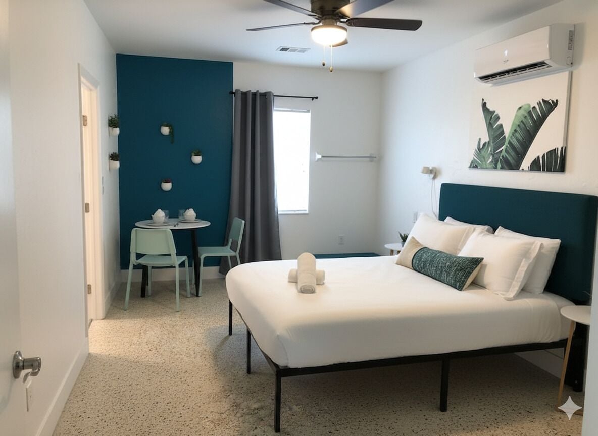A bedroom with a bed, a ceiling fan, an air conditioner, a window with curtains, and a small table with chairs.