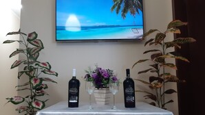 A table with two bottles of wine, two glasses, and a vase of flowers, set against a wall with a TV displaying a beach scene. - You in Paradise in Arraial do Cabo, amazing house facing the beach. Do not miss it !! (Arraial do Cabo)