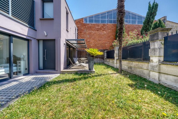 A modern building with a glass entrance, a paved patio, and a grassy area with a palm tree. - La Dépendance, Magnifique maison climatisée avec jardin (Bordeaux)