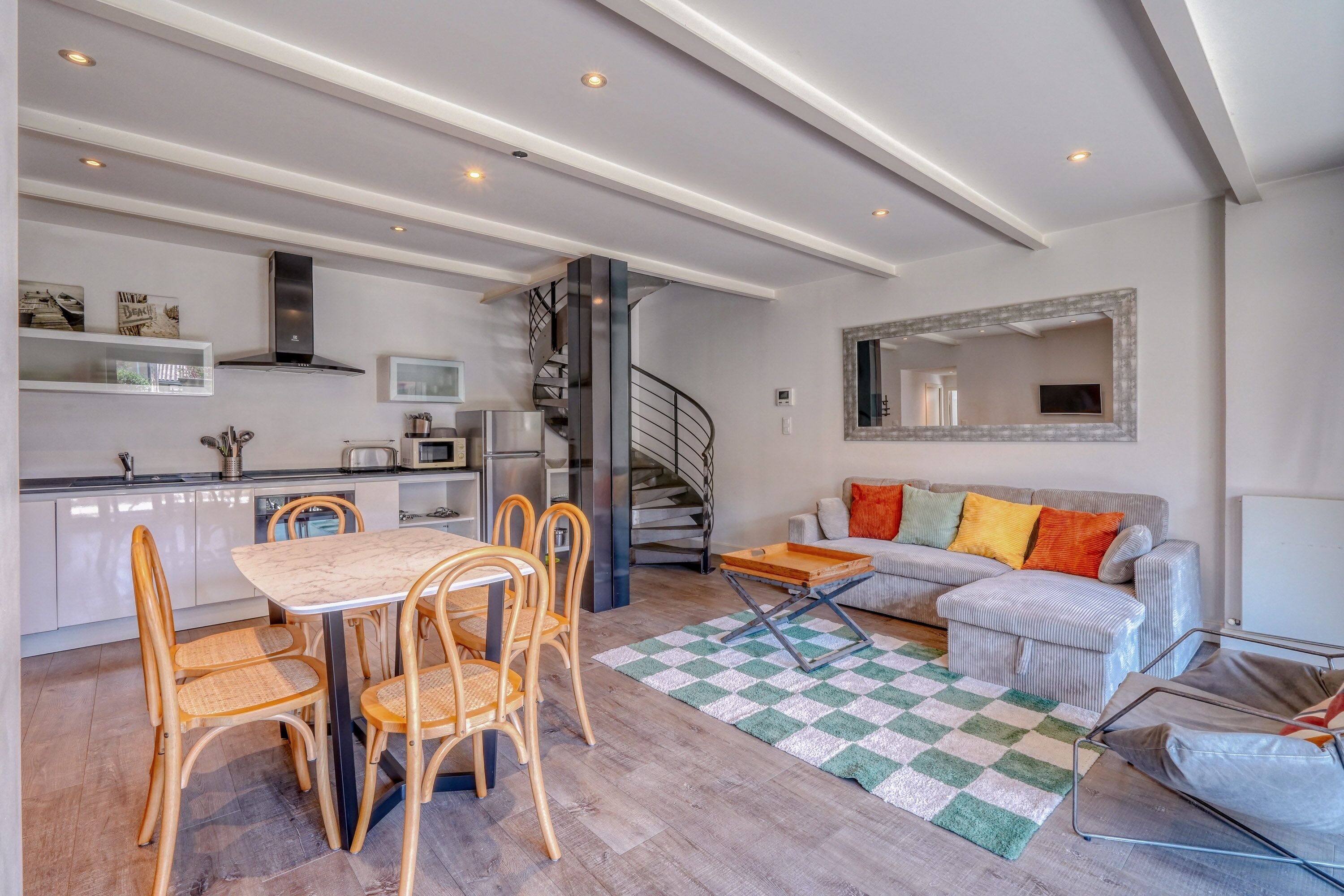 Une cuisine moderne dotée d’appareils électroménagers intégrés, un coin repas avec une table et des chaises en bois, un escalier et un salon avec un canapé et un miroir.