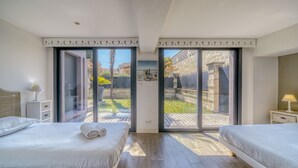 A bedroom with two beds, a nightstand with a lamp, and a view through glass doors to an outdoor area. - La Dépendance, Magnifique maison climatisée avec jardin (Bordeaux)