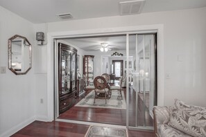 A spacious living room with a dining area, featuring a large mirror, a ceiling fan, and a patterned sofa. - Seas The Day - Steps to Sand! - Monthly Beach Rental (Clearwater Beach)