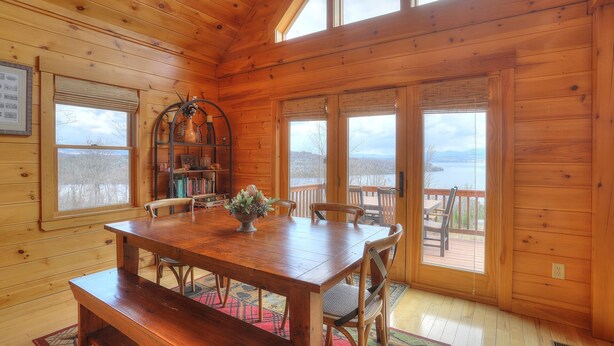 A wooden cabin interior with a dining table, chairs, a bookshelf, and large windows offering a view of a lake and mountains. - 60 West - Beautiful home with STUNNING view of Rangeley Lake (Rangeley)