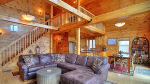 A spacious living area with a large L-shaped sectional sofa, a round coffee table, and a staircase leading to an upper level. - 60 West - Beautiful home with STUNNING view of Rangeley Lake (Rangeley)