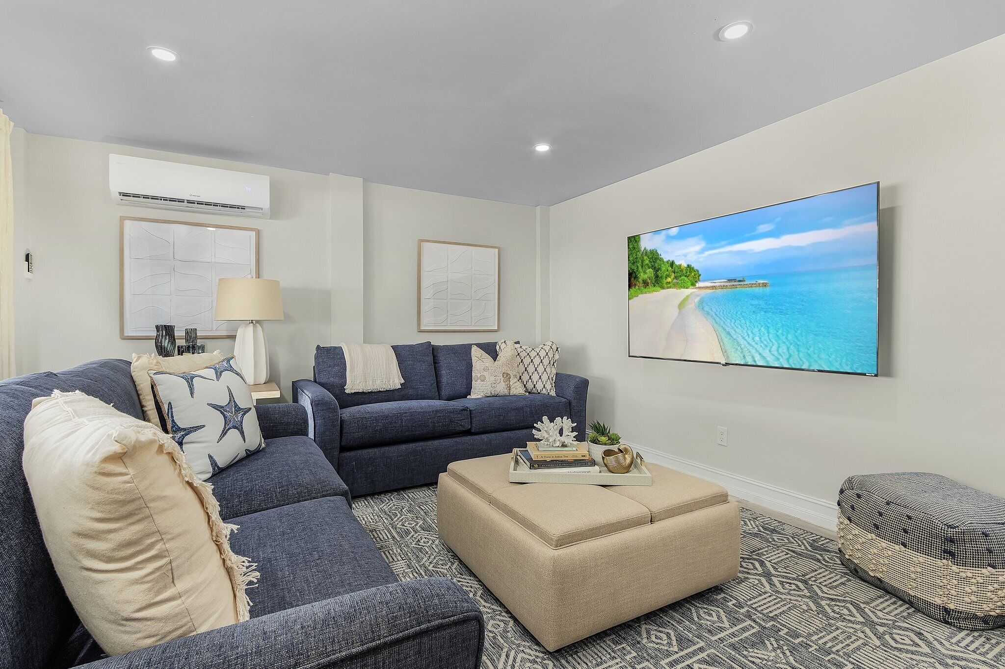 A modern living room with a dark blue sofa, beige cushions, a patterned rug, and a large flat-screen TV displaying a beach scene.