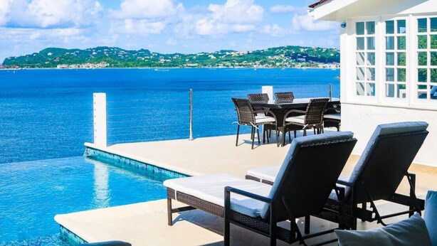Zona junto a la piscina con sillones de descanso y una mesa, con vistas al mar azul turquesa y un paisaje verde distante.